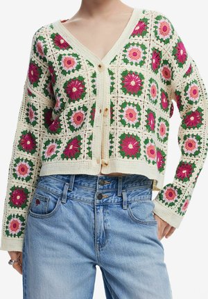 Cardigan en crochet avec des carrés floraux roses et verts sur un jean taille haute bleu clair, devant boutonné et encolure en V.