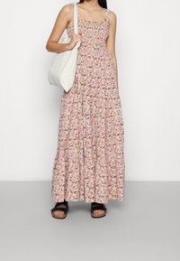 Robe longue rose clair à motif floral, dotée d'un corsage smocké, d'un décolleté carré et de bretelles épaisses. Portée avec un sac fourre-tout beige et des sandales.