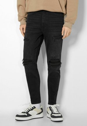 Person wearing black ripped jeans paired with black, white, and beige sneakers, and a beige sweatshirt with visible sleeves.