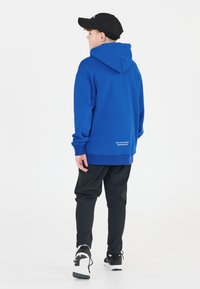 Sweatshirt à capuche bleu vif, avec une coupe décontractée et des poignets côtelés. Associé à un pantalon noir et des sneakers avec des accents blancs.