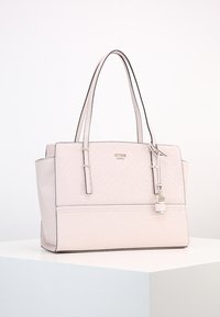 Light pink structured Guess tote bag with dual handles, subtle logo pattern, and hanging small padlock charm on a white surface.