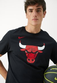 Black cotton T-shirt featuring a large red and white Chicago Bulls logo on the front. Model holds a black and yellow basketball.