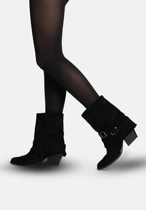 Gambe con calze nere coprenti e stivali di camoscio neri a metà polpaccio con tacchi a blocco e cinturini con fibbia.