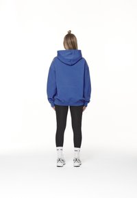 Osoba s dlouhými vlasy svázanými do buně, oblečená v modré mikině, černých legínách, bílých ponožkách a bílých teniskách, stojící zády k nám.