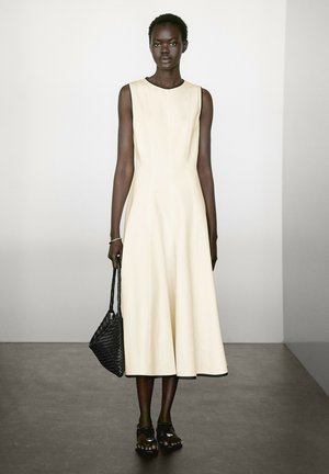 Cream sleeveless dress with a flared skirt design, featuring contrasting black trim. Model holding a black woven handbag, paired with sandals.