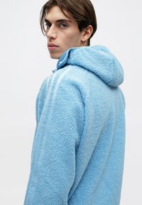 Giovane con capelli corti castani che indossa una giacca in pile azzurro chiaro con cappuccio, che guarda oltre la spalla contro uno sfondo neutro.