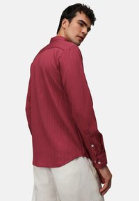 Camicia a maniche lunghe a righe rosse, con colletto e polsini a bottoni, realizzata in tessuto texturizzato e con orlo curvo, indossata sopra pantaloni chiari.