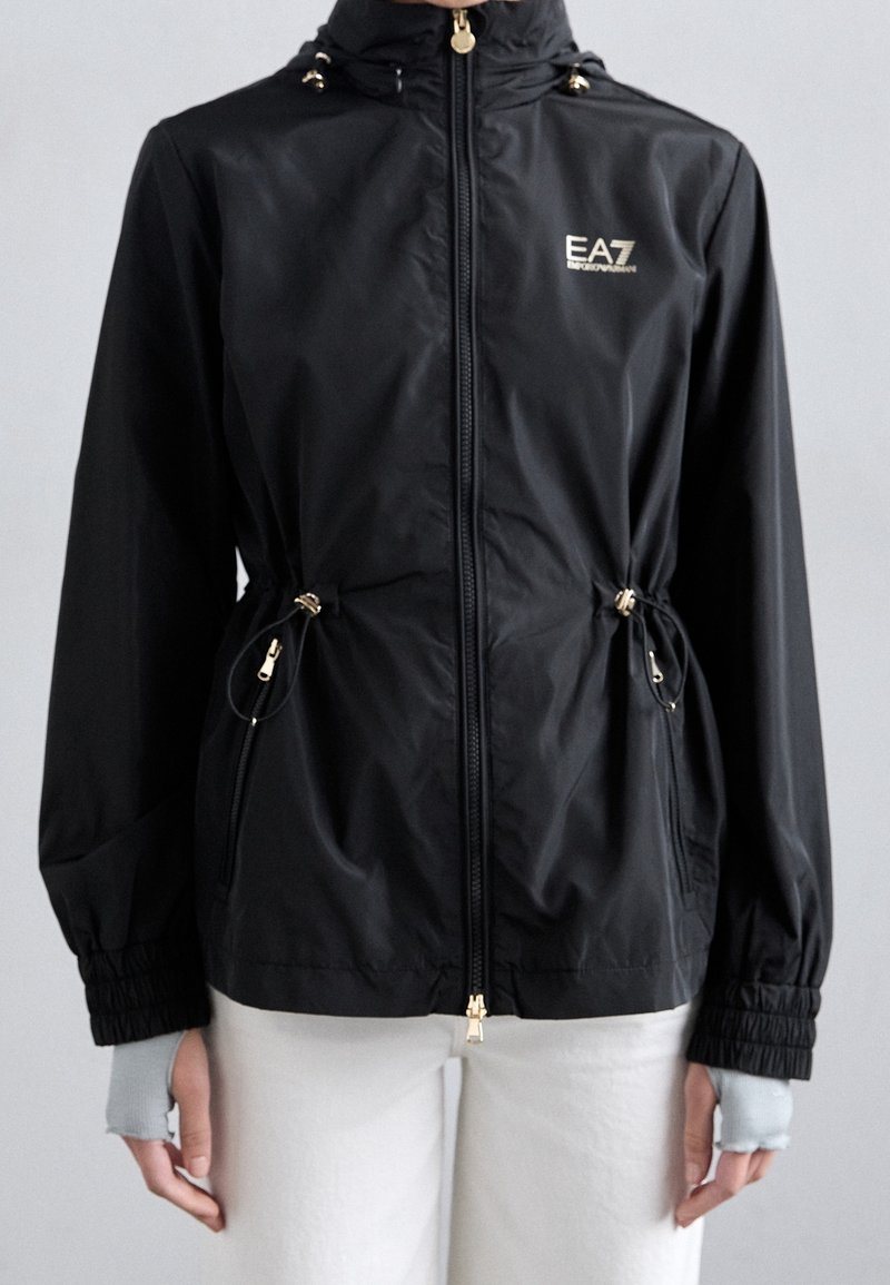 Giacca nera con cerniera e coulisse in vita, con logo "EA7 Emporio Armani" sul petto, indossata con pantaloni bianchi e guanti senza dita.