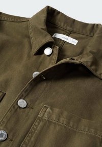 Surchemise vert olive avec boutons-pression argentés, col pointu, poche poitrine et étiquette de marque à l'intérieur du col.