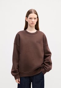Brown oversized sweatshirt with ribbed crew neckline and embroidered logo detail on chest, paired with dark denim trousers.