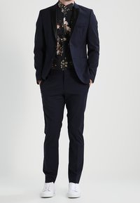 Marineblazer met zwarte satijnen revers over een bloemrijk overhemd, gecombineerd met bijpassende broek en witte sneakers. Gladde stof en een nauwsluitende pasvorm.