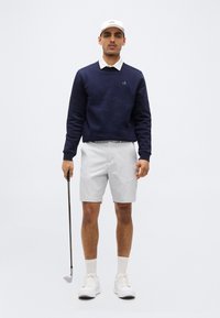 Άνδρας που φοράει ναυτικό φούτερ, λευκό πουκάμισο με γιακά, ανοιχτό γκρι σορτς, λευκό καπέλο και παπούτσια, κρατώντας ένα γκολφ ρόπαλο, στέκεται μπροστά σε απλό φόντο.