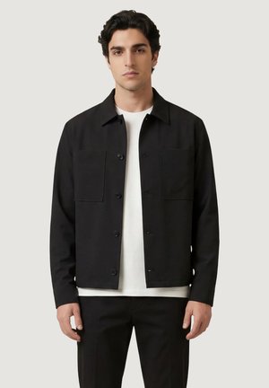 Giovane uomo che indossa una giacca nera con bottoni sopra una camicia bianca e pantaloni neri, in piedi davanti a uno sfondo chiaro e semplice.