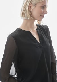 Svart blus med v-ringning och lätta, genomskinliga ärmar. Har en subtil glans och texturerad knappdetalj längs halsringningen.