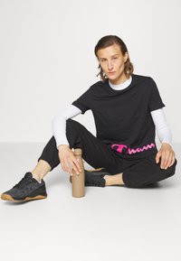 Una camiseta negra con el logo en rosa de "Champion", superpuesta sobre una camisa de manga larga blanca. Joggers negros ajustados y zapatillas negras. Botella de agua beige.