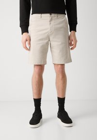 Beige shorts made of cotton, featuring a flat front, side pockets, and a hem just above the knee. Worn with black socks and sneakers.