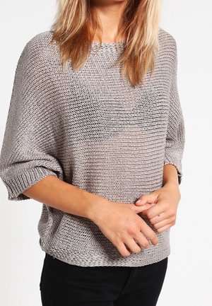 Femme portant un pull en maille gris ample à manches trois-quarts et un pantalon noir, debout, mains jointes.