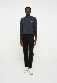Marineblauwe crewneck sweatshirt met wit logo, gecombineerd met zwarte rechte jeans en witte sneakers met beige accenten.