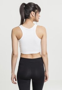 Biely skrátený tank top s hlbokým výstrihom a širokými ramienkami, spárovaný s vysoko pásovými čiernymi legínami, z pohľadu zozadu.