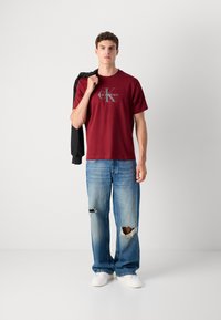 Mladic u bordo Calvin Klein majici, poderanim plavim farmerkama, bijelim tenisicama, drži crnu jaknu preko ramena, nasuprot jednobrojnoj pozadini.