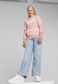 Sudadera con capucha rosa con el logo blanco de "PUMA", combinada con unos vaqueros de pierna ancha de color azul claro y zapatillas de plataforma blancas, de pie sobre una superficie blanca.