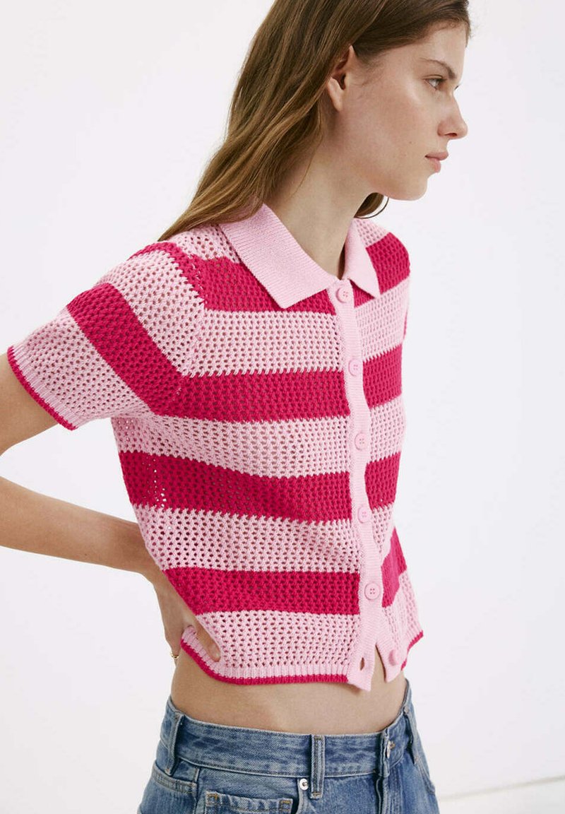 Jeune femme portant un haut en maille à manches courtes rayé rose et rouge avec des boutons, associé à un jean en denim bleu, debout de profil, main sur la hanche.