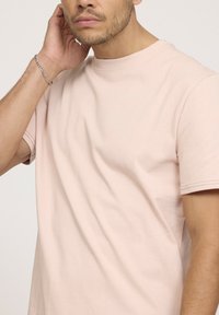 T-shirt en coton rose clair avec un col rond et des manches courtes. Texture lisse, coupe décontractée. Bracelet en chaîne argent sur le poignet.