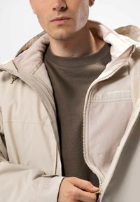 Beige vedenkestävä takki, jossa on fleece-vuori, vetoketjullinen etuosa ja säädettävä huppu; päällä ruskea huppari. Huomattava brändilogo näkyvissä.