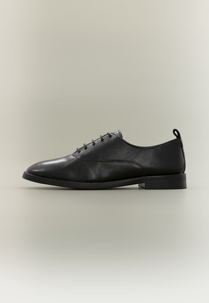 Chaussure de ville en cuir noir avec petit talon, lacets à l’avant et languette de préhension sur fond neutre, vue de côté.