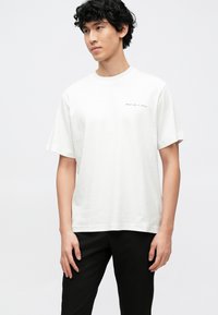 Λευκό βαμβακερό μπλουζάκι με στρογγυλή λαιμόκοψη και κοντά μανίκια. Διαθέτει μικρό μαύρο κείμενο στο στήθος. Συνδυάζεται με μαύρο παντελόνι.