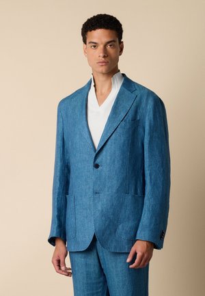 Homme portant une veste de costume bleue et un pantalon assorti sur un t-shirt blanc à col en V, debout devant un fond beige uni.