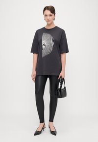 Crni oversized T-shirt s bijelim grafičkim motivom sunca; uparen s crnim tajicama, crnim štiklama s čavlima i malom crnom torbom s remenom.