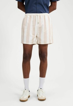 Persona che indossa pantaloncini beige e bianchi a righe verticali, calzini bianchi, scarpe da ginnastica bianche e una camicia blu scuro infilata dentro.