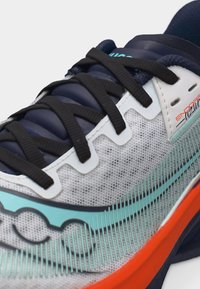 Κοντινό πλάνο αθλητικού παπουτσιού με μαύρα κορδόνια, επάνω μέρος από λευκό και γκρι δίχτυ, σχέδιο σε ανοιχτό μπλε και φωτεινή πορτοκαλί σόλα.