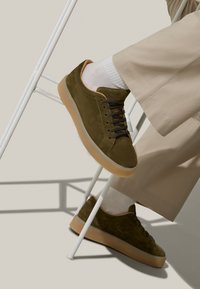 Sneaker in pelle scamosciata verde oliva con suola in gomma, dettagli beige, punta rotonda e lacci neri. Calze bianche visibili sopra pantaloni color sabbia.