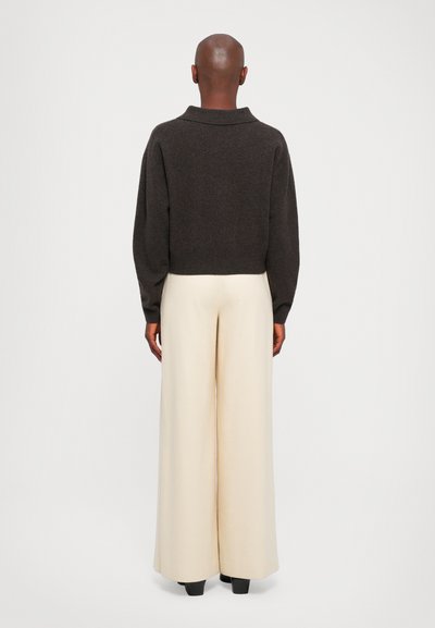 Une personne se tenant de dos, portant un pull marron foncé, un pantalon large crème et des chaussures noires, sur un fond clair et uni.