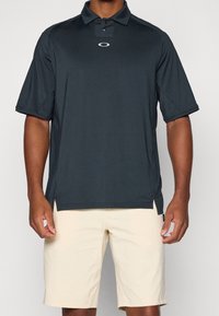 Polo shirt gris foncé avec une patte de boutonnage à deux boutons et des fentes sur les côtés. Associé à un short beige clair, avec une texture lisse et une coupe décontractée.