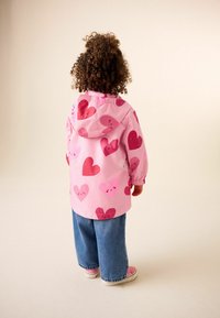 Růžová nepromokavá bunda s velkými červenými a růžovými srdcovými vzory, kapucí, elastickými manžetami a měkkou texturou, doplněná světle modrými džínovými kalhotami a teniskami.