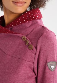 Primo piano di una persona che indossa un maglione bordeaux con un ampio colletto, dotato di tre bottoni decorativi in legno e una fodera interna a motivo.