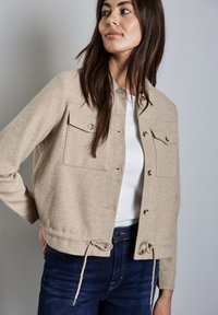 Beige cropped bunda z vlněné směsi se dv�ěma předními kapsami s klopami, zdobená zlatými knoflíky a stahovacími lemy. Nosená přes bílý top.