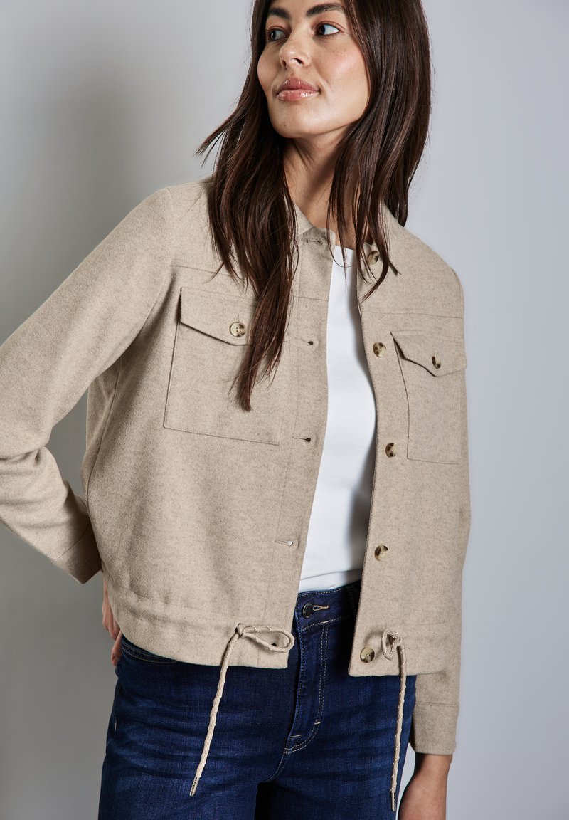 Beige cropped bunda z vlněné směsi se dvěma předními kapsami s klopami, zdobená zlatými knoflíky a stahovacími lemy. Nosená přes bílý top.