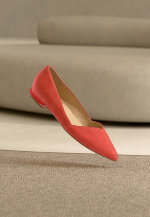 Chaussure plate rouge à bout pointu suspendue au-dessus d'un tapis beige texturé avec un canapé beige incurvé en arrière-plan.