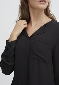 Svart bluse med V-hals, kneppede ermer og en enkelt brystlomme. Laget av et glatt, lett stoff med en litt løs passform.