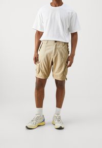 Beige cargo shorts with multiple side pockets, made of lightweight fabric. Paired with a plain white t-shirt and athletic sneakers.