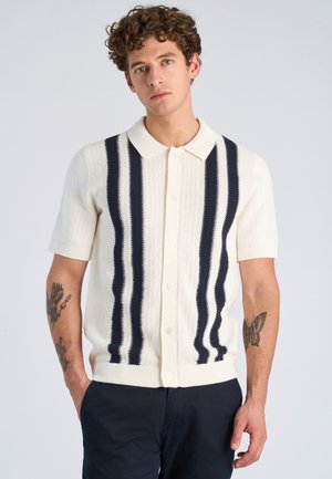 Młody mężczyzna z kręconymi włosami, ubrany w biały sweter na guziki z krótkim rękawem i czarnymi pionowymi paskami oraz ciemne spodnie, stoi z rękami w kieszeniach.