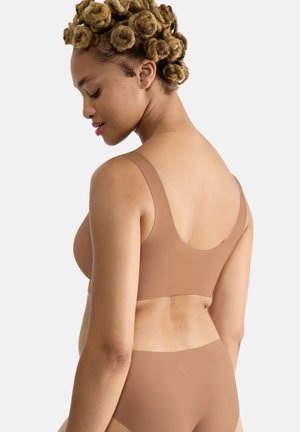 Frau mit kurzen, gelockten Haaren, die einen nahtlosen beigen BH und passende Unterwäsche trägt, von hinten vor einfarbigem Hintergrund gezeigt.
