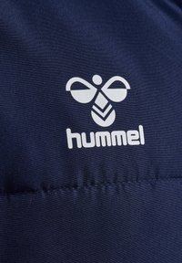 Mørkeblå stof med en glat tekstur, der prydes af et hvidt logo af to vinger over et bladmønster og brandnavnet "hummel" i fed skrift nedenfor.