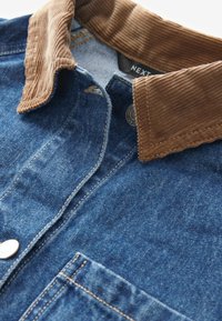 Koszula dżinsowa z brązowym kołnierzem z sztruksu, przednią kieszenią, metalowym guzikiem oraz widocznymi detalami przeszycia na teksturowanej niebieskiej tkaninie.