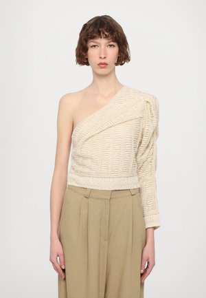 Asymmetrisk beige topp med teksturert strikkemønster, én lang arm, ribbestrikket kant, kombinert med høy midje beige bukser.