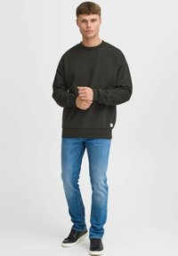 Mørkegrønn genser med rund hals, ribbestrikkede mansjetter og nederkant, kombinert med lyseblå jeans og svarte joggesko. Avslappet, løs passform.